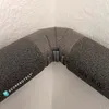 A close-up view of a gray pipe insulation corner with clips, attached to a textured white wall. Produktbild-Vorschau