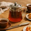 A glass teapot filled with tea, accompanied by a cup of tea and croissants on a wooden table. Podgląd zdjęcia produktu