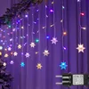 A string of colorful LED snowflake lights hanging against a purple curtain, with a Christmas tree visible on the left. Náhled obrázku produktu