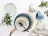 A collection of ceramic plates, bowls, and mugs arranged on a white surface with some greenery. Náhľad obrázku produktu