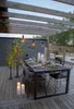 An outdoor dining table set up under a wooden pergola, decorated with string lights and candles. Produktbild-Vorschau