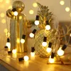 A string of decorative lights is draped around golden decorative objects, including a cactus and a pineapple, on a wooden surface. Náhled obrázku produktu