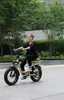 A woman rides an electric bicycle on a paved path surrounded by greenery and modern buildings. Produktbild-Vorschau