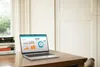 A laptop displaying charts and graphs is placed on a wooden desk next to a stack of books, with a window and curtains in the background. Voir les images du produit