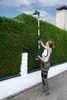 A woman is using a telescopic hedge trimmer to trim a tall hedge. Produktbild-Vorschau