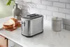 A silver toaster sits on a white countertop next to sliced bread, a vase, and a decorative container. Anteprima dell’immagine prodotto