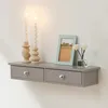 A gray floating shelf with two drawers is mounted on a white wall. It is decorated with two white twisted candles on wooden candle holders, a framed picture of a landscape, and a small glass bottle with reeds. Podgląd zdjęcia produktu