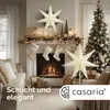 A cozy living room decorated for Christmas, featuring a fireplace, stockings, a Christmas tree, and decorative stars. Produktbild-Vorschau