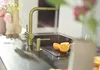A modern kitchen sink area with a gold faucet, soap dispenser, and sliced oranges on a cutting board. Produktbild-Vorschau