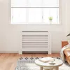 A white radiator cover is shown in a living room setting, placed in front of a window with blinds. A small potted plant sits on the windowsill above the cover. A round coffee table with a book and a cup of coffee is in the foreground, and part of a brown l Podgląd zdjęcia produktu