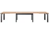 A long, extendable dining table with a wooden top and black metal legs, shown against a white background. Produktbild-Vorschau