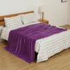 A purple blanket is draped over a bed with checkered bedding. A bedside table with a lamp is visible. Produktbild-Vorschau