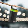 A black portable speaker with a colored light ring is hanging from a tree branch outdoors, with blurred lights in the background. Voir les images du produit