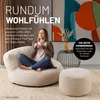 A woman is relaxing on a large beanbag chair with a cup of coffee, next to a small ottoman. The scene is set in a modern living room with abstract art on the wall. Podgląd zdjęcia produktu