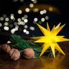 A yellow star decoration with pine needles and walnuts on a dark surface, with bokeh lights in the background. Produktbild-Vorschau
