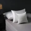 Three white silk pillowcases are arranged on a bed with a bedside table and lamp in the background. Podgląd zdjęcia produktu