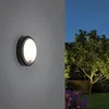An outdoor wall light is mounted on a textured gray wall, with a garden scene featuring a tree and flowers visible to the right. Produktbild-Vorschau