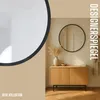 A round black mirror with a metal rim hangs on a wall above a wooden cabinet with two doors. A vase with dried grass and a small bowl are on the cabinet. A bathrobe hangs on a hook to the right of the mirror. Produktbild-Vorschau