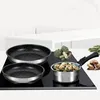 A set of cookware, including two frying pans and a small saucepan with a removable handle, is displayed on a black induction stovetop. Various vegetables and mushrooms are arranged around the cookware. Voir les images du produit