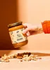 A hand holding a jar of peanut butter with peanuts scattered around the base. Produktbild-Vorschau