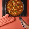 A hand holding a black and silver kitchen utensil over a pan of food on an oven rack, with a pink oven mitt nearby. Voir les images du produit
