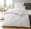 A white comforter is laid out on a bed, with pillows behind it. A small side table with a vase and bowl is to the left. Produktbild-Vorschau