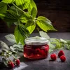 A jar of raspberries and fresh raspberries are arranged with raspberry leaves on a dark surface. Produktbild-Vorschau