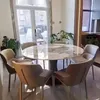 A dining room with a round table and chairs, with plants and a large window in the background. Produktbild-Vorschau