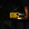 A yellow emergency radio is placed on a shelf with plants and a terracotta pot in the background, emitting a beam of light. Produktbild-Vorschau
