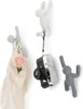 Three wall hooks shaped like figures climbing on a white wall. One hook holds a bag with a flower, another holds a camera, and the third is empty. Náhled obrázku produktu