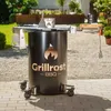 A black steel fire pit with a grill grate and accessories, set up on a paved patio in a garden. Produktbild-Vorschau