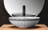 A decorative washbasin with a black faucet is placed on a wooden surface in front of a large round mirror. Produktbild-Vorschau