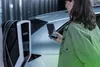 A woman is holding a smartphone and standing next to an electric vehicle charging station. Produktbild-Vorschau