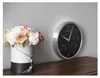A wall clock with a black face and silver frame is mounted on a light gray wall, next to a vase of flowers. Podgląd zdjęcia produktu