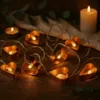 A string of heart-shaped LED lights is arranged on a wooden surface, with candles and greenery in the background. Produktbild-Vorschau