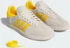 A pair of white and yellow Adidas Samba sneakers with a yellow shoelace coiled on the side. Produktbild-Vorschau