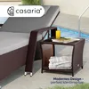 A brown polyrattan side table with a glass top and a shelf, placed next to a lounge chair by a swimming pool. A glass of yellow liquid and magazines are on the table, and towels are on the shelf. Anteprima dell’immagine prodotto