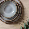A stack of beige plates and bowls on a wooden surface, with two small cards listing menu items and a sprig of eucalyptus. Produktbild-Vorschau