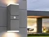 A modern outdoor wall light with a motion sensor is mounted on a gray building facade, with a blurred view of a house interior and lawn in the background. Anteprima dell’immagine prodotto