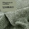 A close-up of a textured sage green fabric, likely a pillowcase, with care instructions and symbols overlaid on a light green background. Produktbild-Vorschau