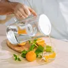 A person is pouring water from a pitcher into a glass, with oranges and mint leaves on a wooden board. Anteprima dell’immagine prodotto