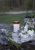 A white LED candle with a decorative band sits on a stone surface, surrounded by ivy and a white lily. Produktbild-Vorschau