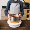 A person wearing an apron is holding a cake ring around a cake on a cake stand. Podgląd zdjęcia produktu