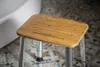 A close-up of a bamboo shower seat with silver legs, placed on a patterned tiled floor next to a white bathtub. Anteprima dell’immagine prodotto