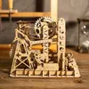 A wooden mechanical model, possibly a marble run or automaton, is displayed on a wooden surface. An hourglass is visible in the background. Voir les images du produit