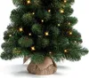 A close-up view of the bottom portion of an artificial Christmas tree, decorated with small LED lights and a burlap base. Náhled obrázku produktu