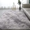 A fluffy gray rug is placed on a gray floor in a living room setting, with a sofa and a small side table with a plant visible. Náhled obrázku produktu