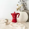 A red Moka pot and two small beige cups are arranged on a light-colored surface with draped fabric. In the background, a large white vase holds dried flowers. Anteprima dell’immagine prodotto