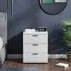 A white nightstand with three drawers is placed next to a bed in a bedroom. There are books and a digital clock on top of the nightstand, and a plant in a basket to the right. The wall is dark blue, and the floor is wooden. Podgląd zdjęcia produktu