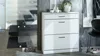 A white shoe cabinet with two drawers is placed in a hallway next to a modern staircase. Decorative items and shoes are visible. Produktbild-Vorschau